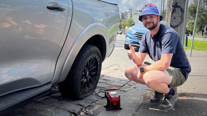A man in a bucket hat squats next to a large ute using an electric pump on its flat tyre. 