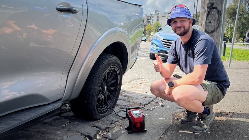 A man in a bucket hat squats next to a large ute using an electric pump on its flat tyre. 