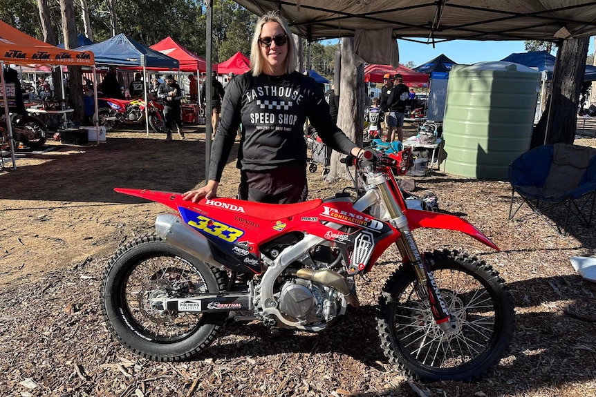 A women in motorbike leathers and sunglasses stands with a red motorbike. 