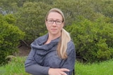 A woman wearing a grey shawl and dark-rimmed glasses standing in front of some shrubby trees