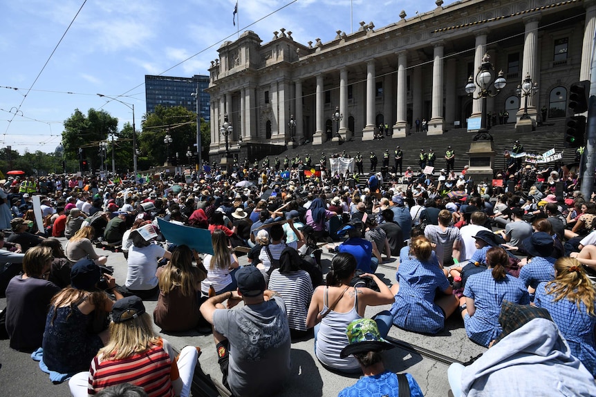 Hundreds of students are seen sitting down in front of Victorian Parliament. The sky is blue.