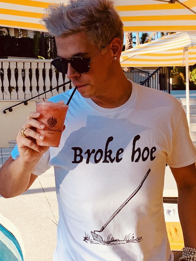 Yiannopoulos sips a drink wearing a white shirt saying "broke hoe" with an image of a garden hoe.
