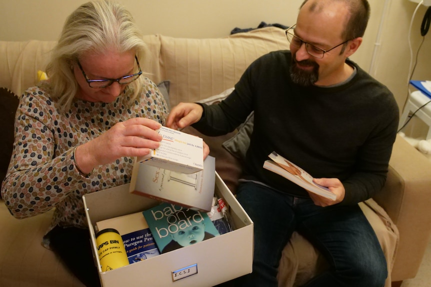 A man and a woman look at books and medication boxes inside a cardboard box