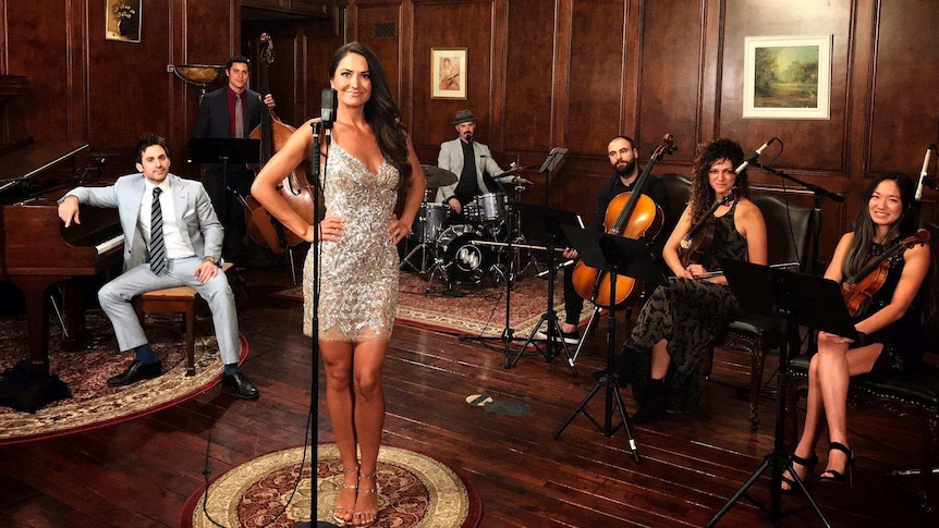 Postmodern Jukebox performers pose in a room with wooden floor and walls with instruments.