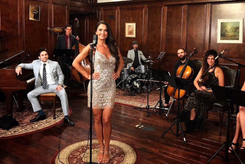 Postmodern Jukebox performers pose in a room with wooden floor and walls with instruments.
