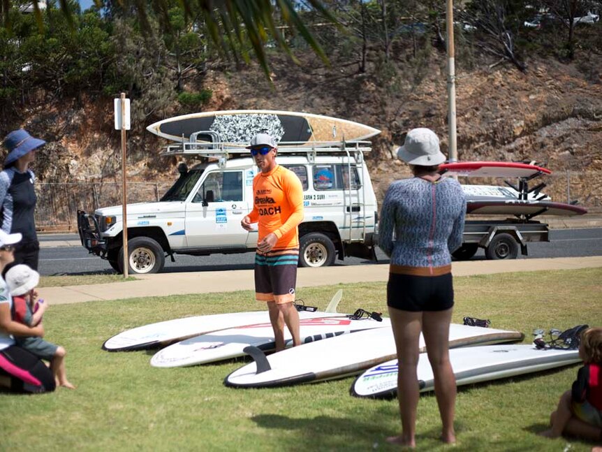 A man in a fluro orange rash shirt stands among paddle boards, instructing a group of mums