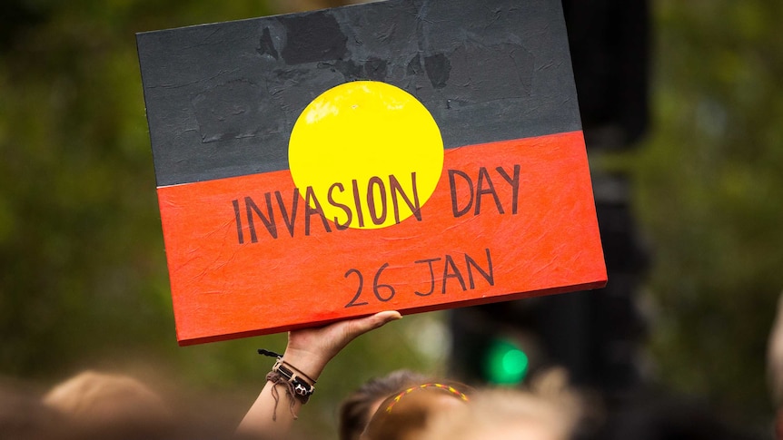 A person holds an Invasion Day sign with the Aboriginal flag at a protest.