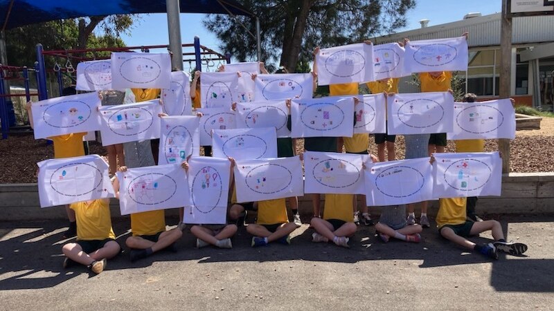 Primary school kids holding up decorated pillowcases in front of a playground.  