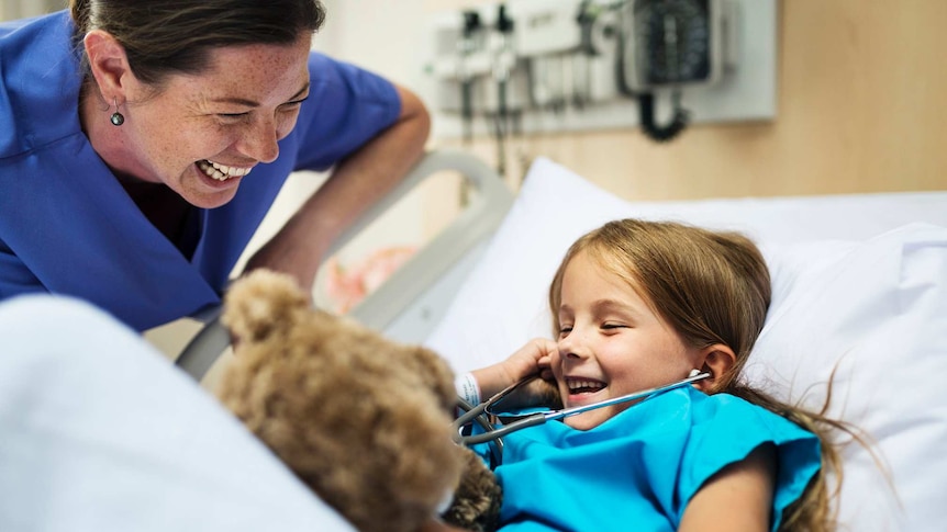 A laughing girl in a hospital bed with a teddy bear