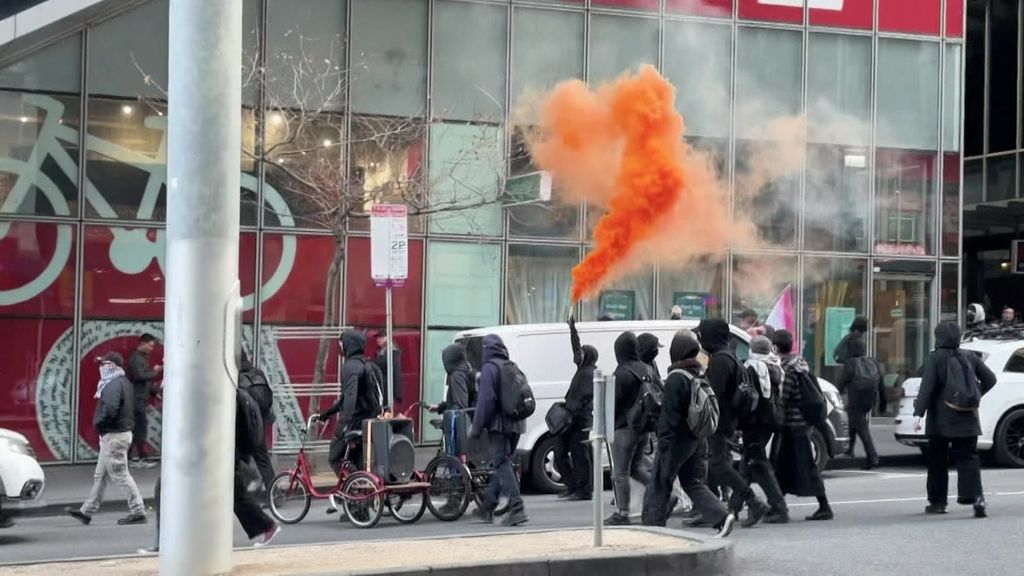 A person dressed in black amid a group of protesters holds a flare oozing orange gas into the air.