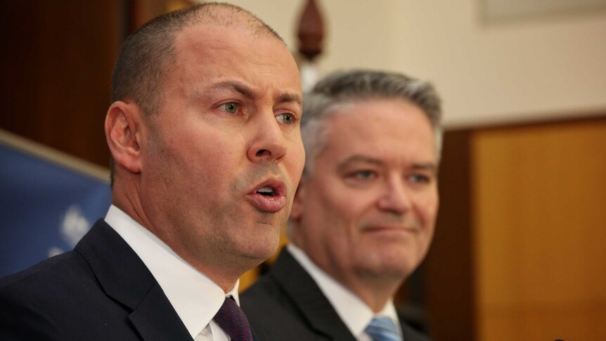 Josh Frydenberg's lips are pursed as he speaks. In the background, somewhat out of focus, Mathias Corman smirks.