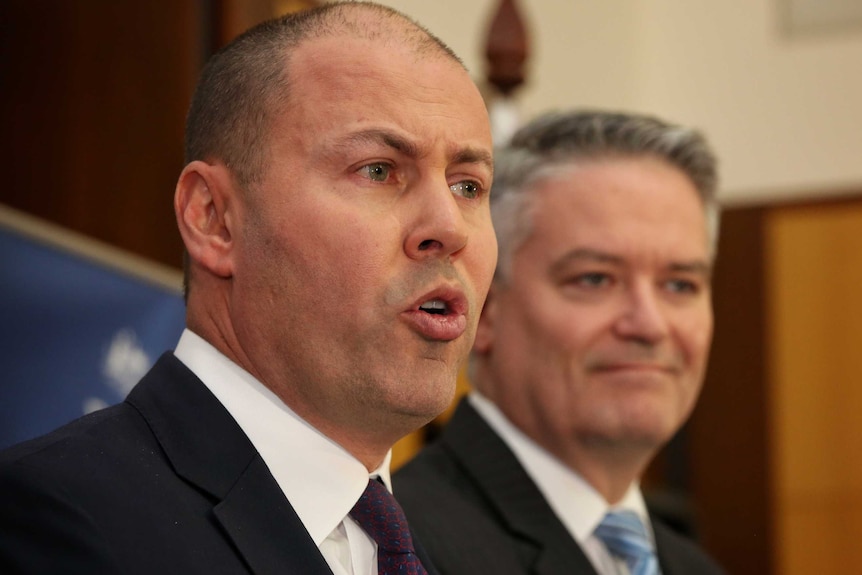 Josh Frydenberg's lips are pursed as he speaks. In the background, somewhat out of focus, Mathias Corman smirks.