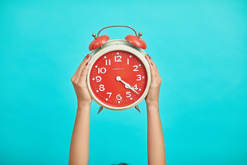 hands hold an orange alarm clock against a blue background