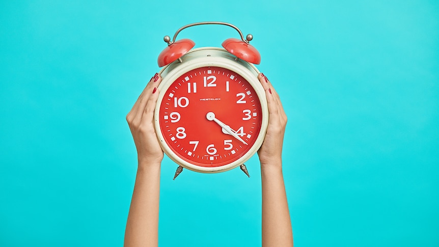 hands hold an orange alarm clock against a blue background