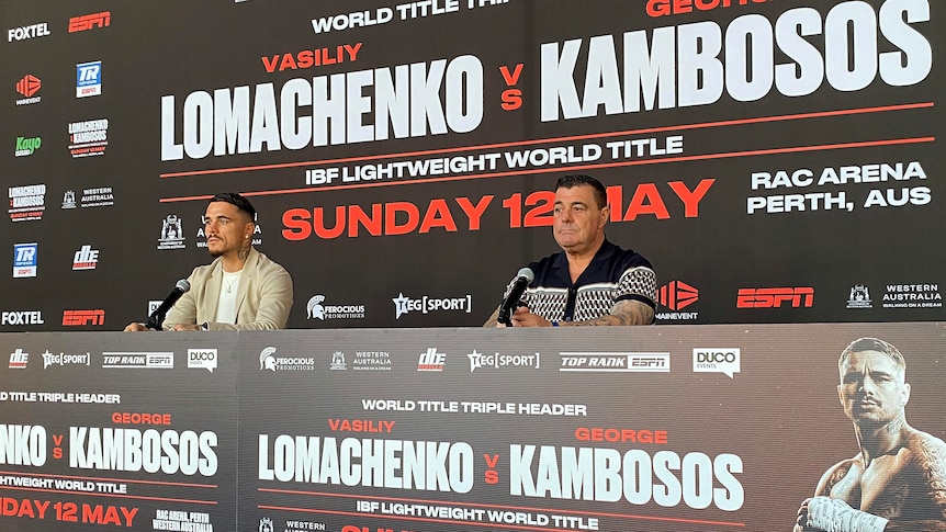 An Australian boxer and his father sit at a desk on a stage with signs 'Vasliy Lomachenko vs George Kambosos'.