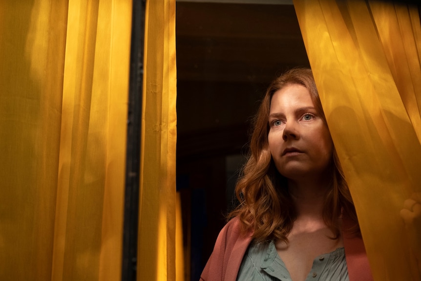 Film still of Amy Adams as Anna cast in shadow looking out the window through yellow curtains in The Woman in the Window