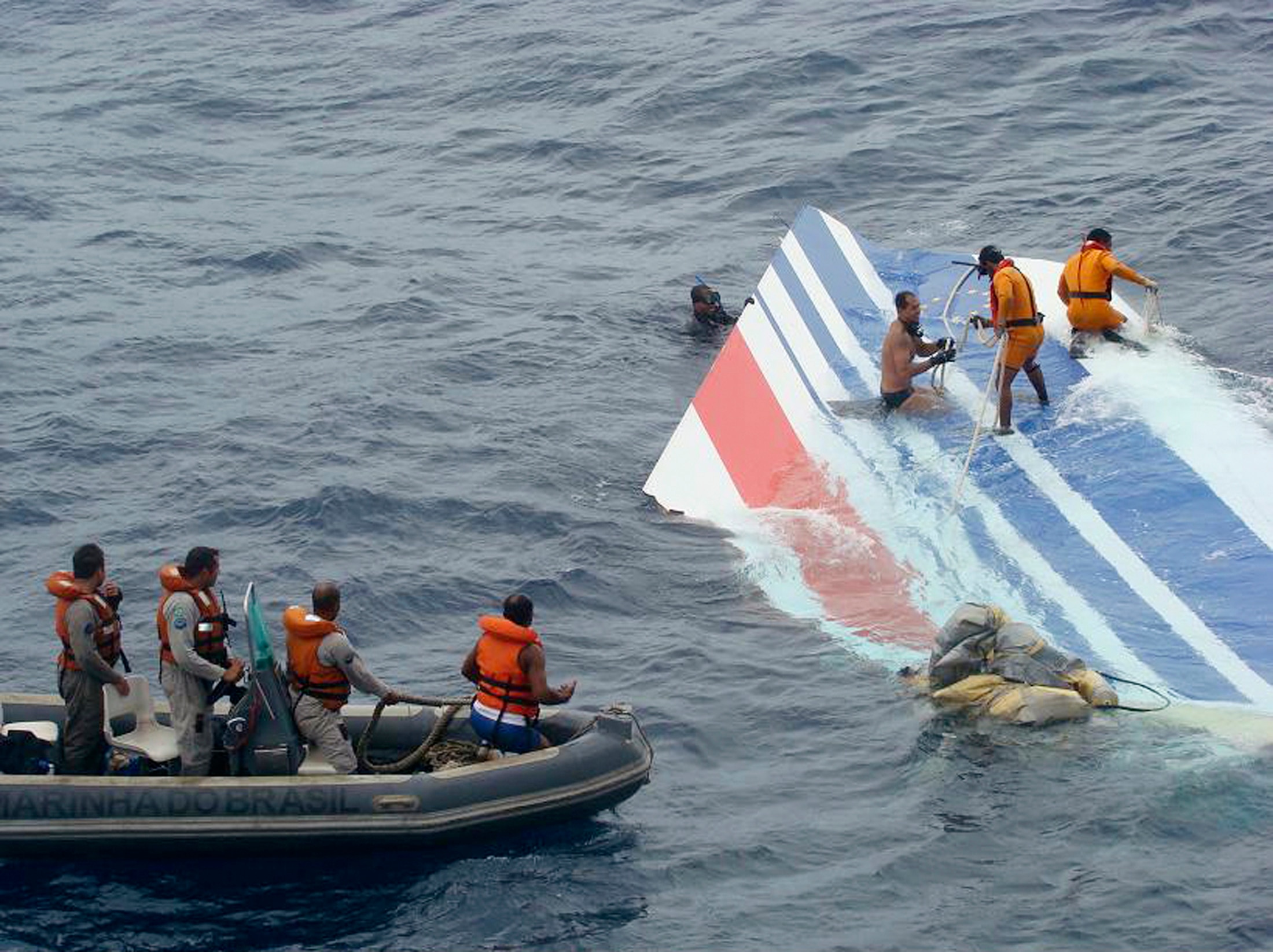 People stand on top of a white, blue and red piece of plane debris in the water while others watch from a rubber boat.