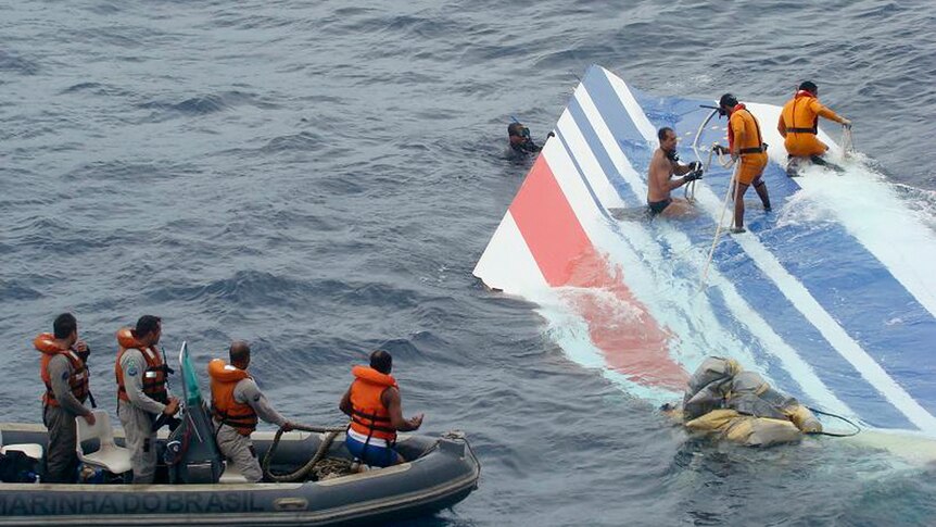 People stand on top of a white, blue and red piece of plane debris in the water while others watch from a rubber boat.