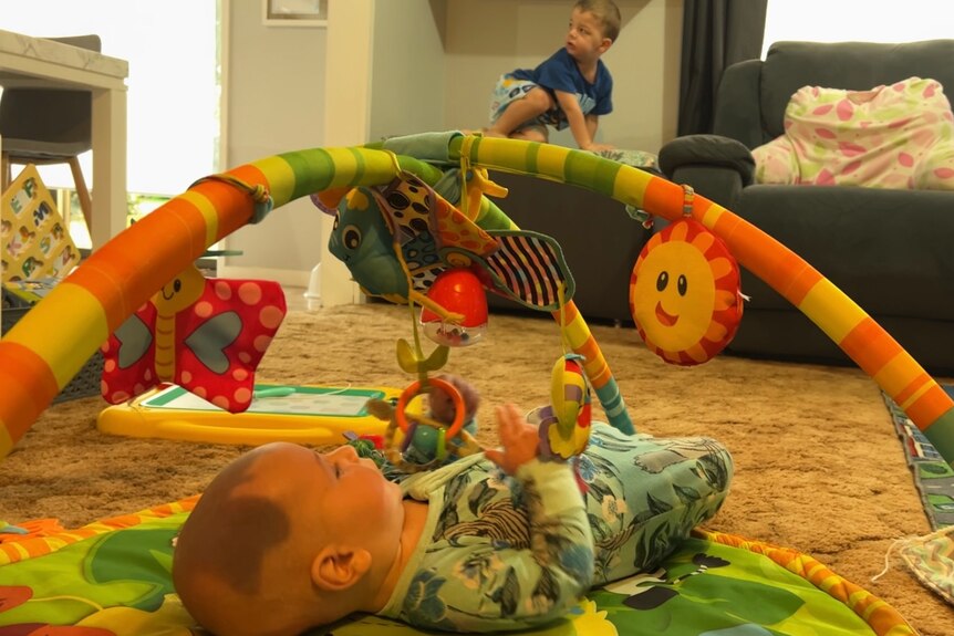 A baby plays with a mobile while a toddler climbs on a couch behind him