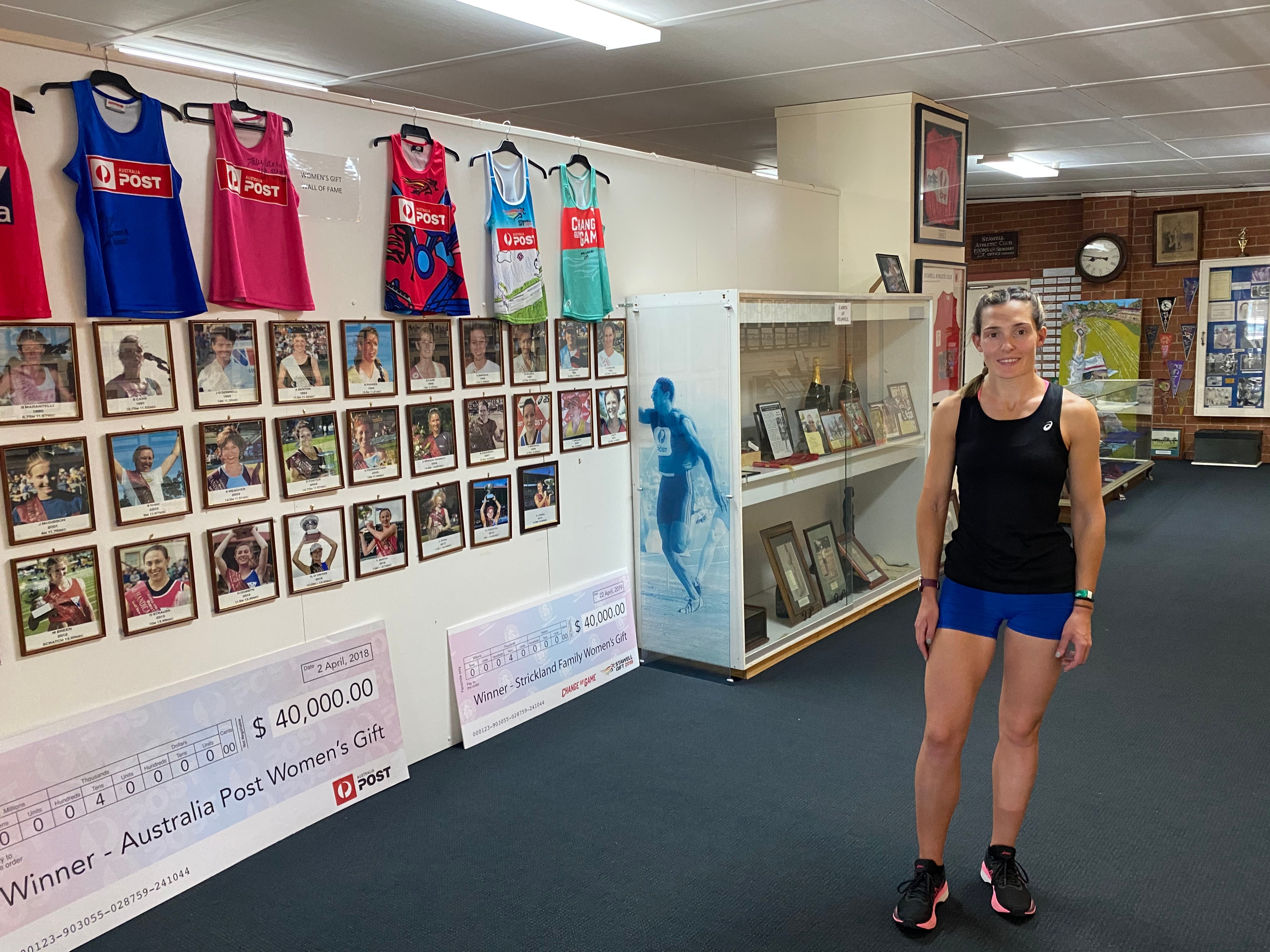 A young woman stands in a room with pictures of former sportsmen all over its walls. Giant novelty cheques are on the floor.