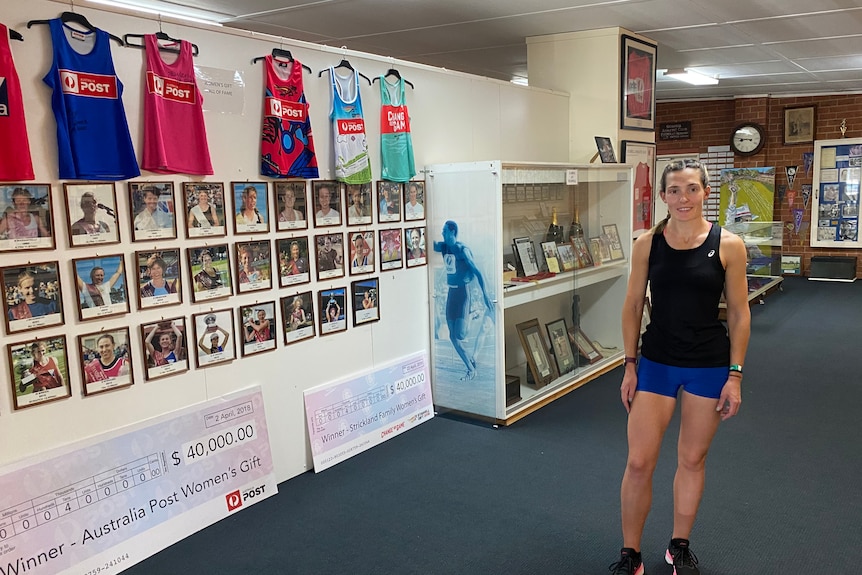 A young woman stands in a room with pictures of former sportsmen all over its walls. Giant novelty cheques are on the floor.