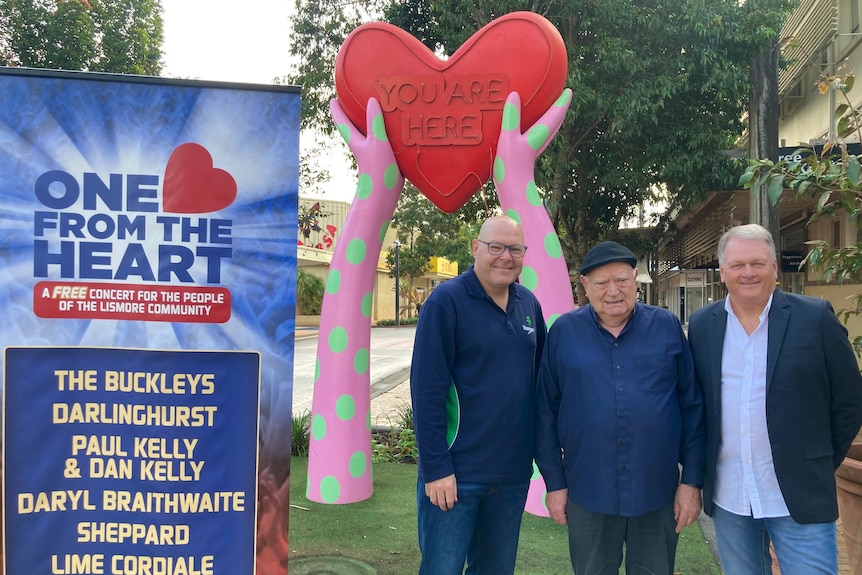 Three men in front of an installation of hands holding up a heart and a poster