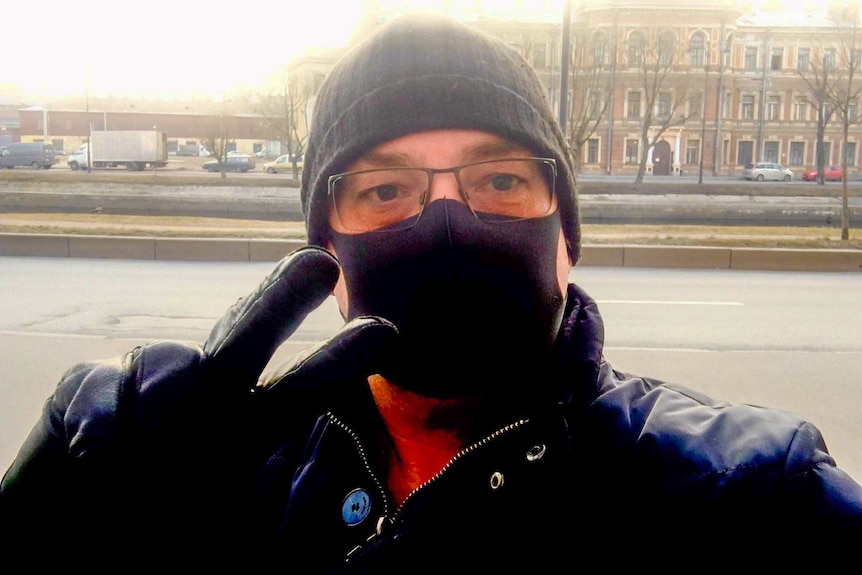 A man in a medical face mask, beanie and sunglasses gives a peace sign to the camera