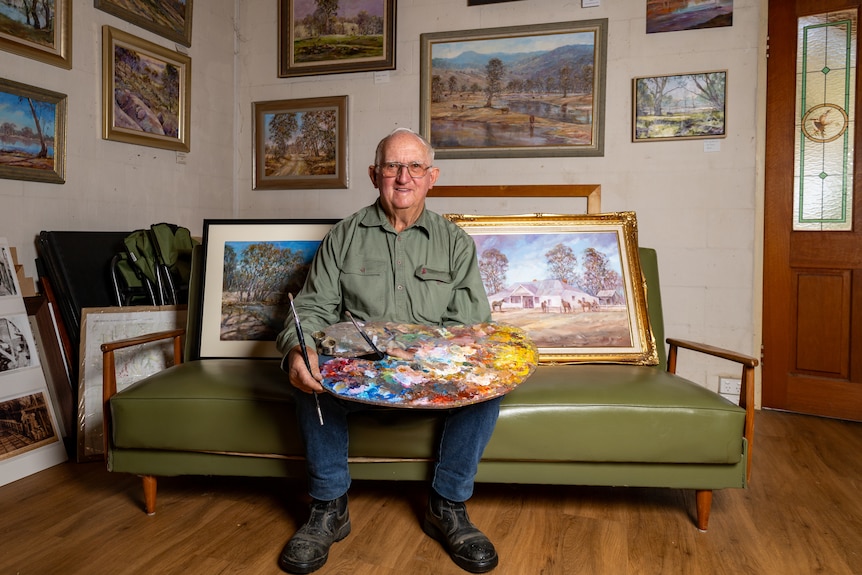 An older man sits on a couch holding a paint palette.