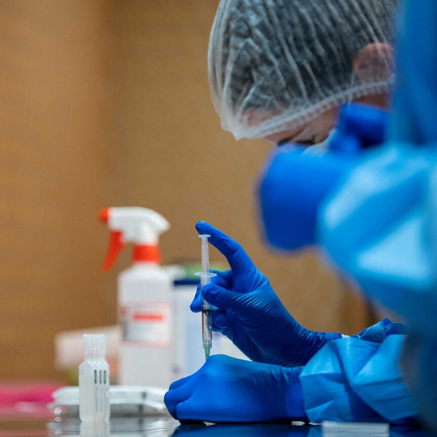A health worker wears blue latex gloves and draws coronavirus vaccine from a vial, on a bench, next to a yellow biohazard tub.