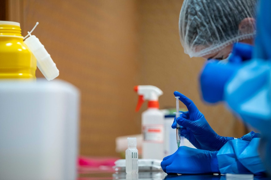 A health worker wears blue latex gloves and draws coronavirus vaccine from a vial, on a bench, next to a yellow biohazard tub.