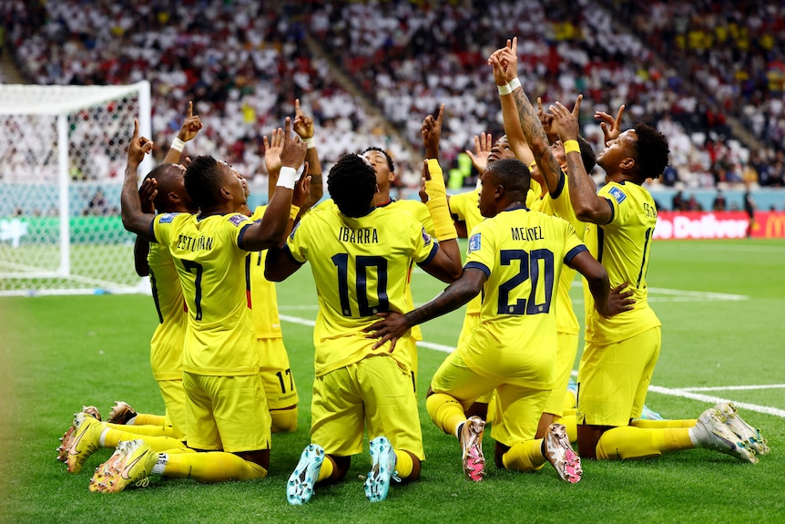 Ecuador players kneel on the field in a circle with their hands raised in celebration.
