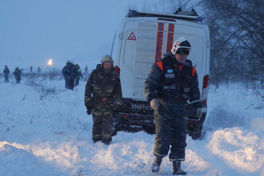 Emergency workers walk through the snow, in front of a white van.