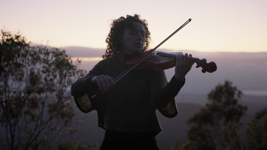 Emily Sheppard plays violin with her eyes closed on a mountain. Behind her is a dusk sky and the tops of gum trees.
