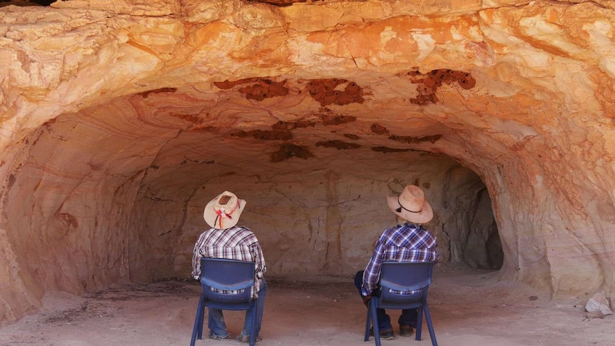 Two men sit in blue plastic chairs facing away from the camera into the cave