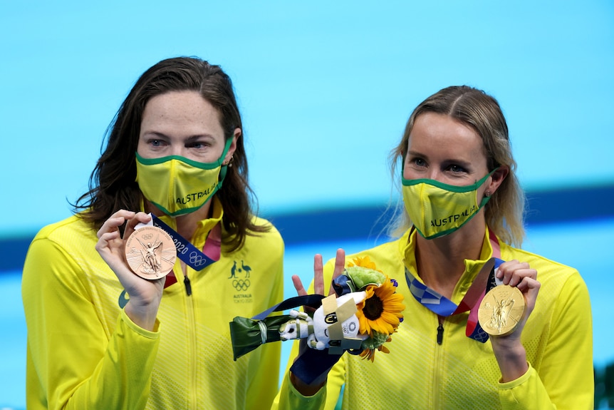 Cate Campbell holds up her bronze medal and Emma McKeon her gold. Both are wearing gold face masks and uniforms