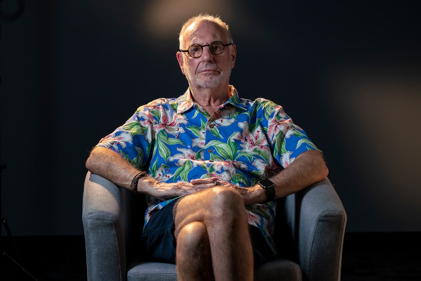 A man in glasses and a Hawaiian shirt sitting on a chair, inside a dark studio. 