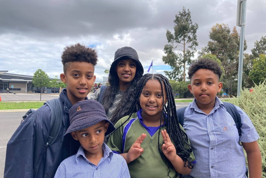 four children wearing school uniforms and a woman standing on a suburban street.