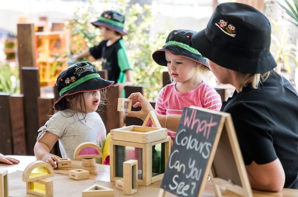 Three young children sitting at a table are shown a block by an adult. A nearby sign reads 'What colours can you see?'.