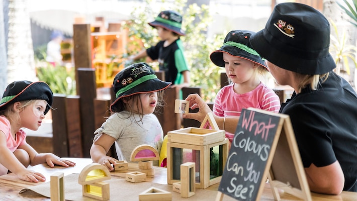 Three young children sitting at a table are shown a block by an adult. A nearby sign reads 'What colours can you see?'.