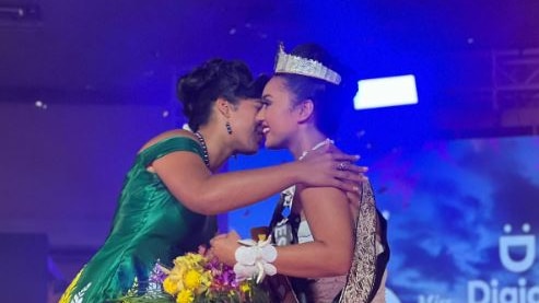 Woman in green dress kisses newly crowned Miss Samoa wearing white dress, holds flowers in room. Contestants stand behind. 