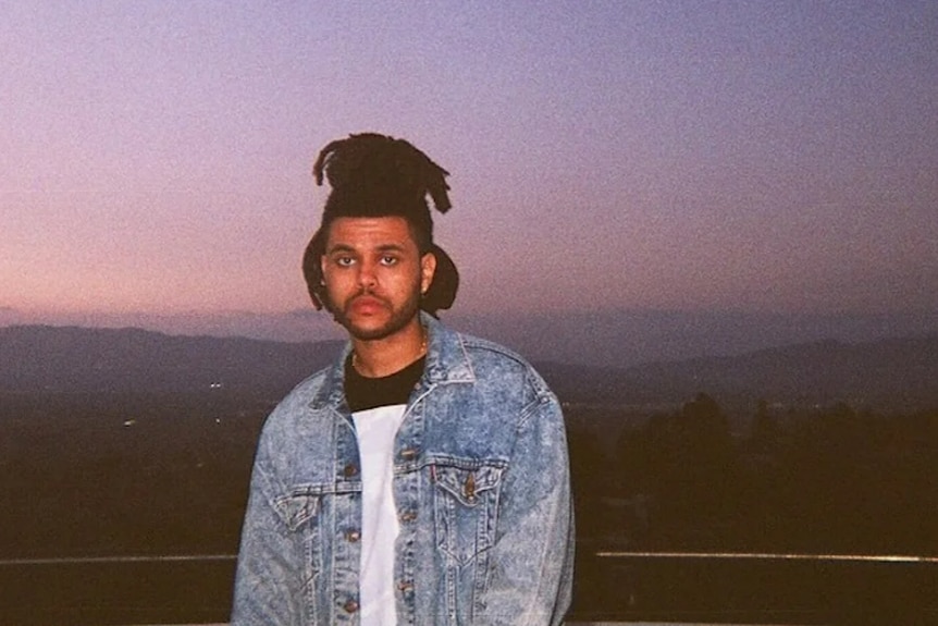 A man wearing a denim jacket poses in front of a pink sunset