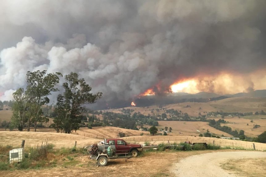 Smoke and flames rise over paddocks as a ute sits on a driveway.