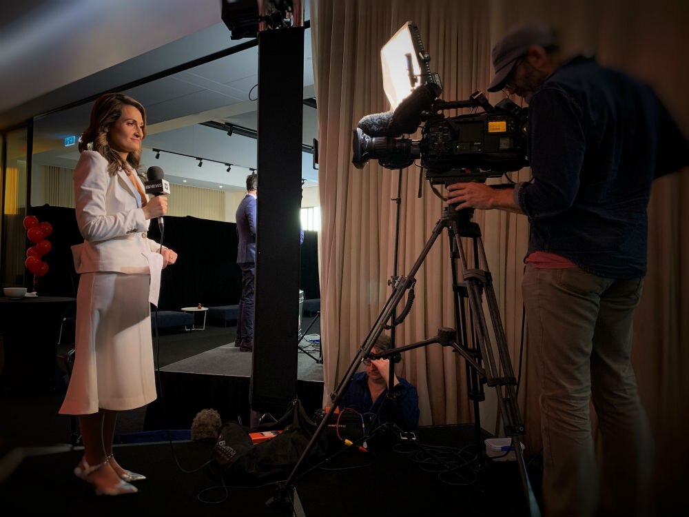 Camera filming Karvelas holding microphone standing on platform.
