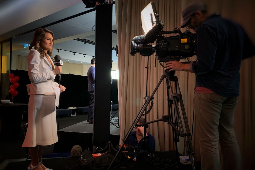 Camera filming Karvelas holding microphone standing on platform.