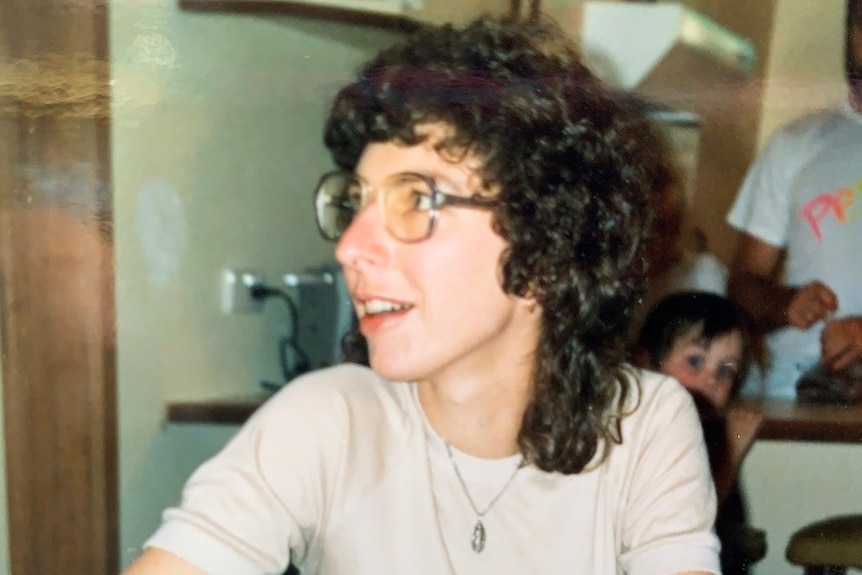 Stephanie Piper with long curly hair and big glasses sits at a kitchen table in what appears to be an old photograph.