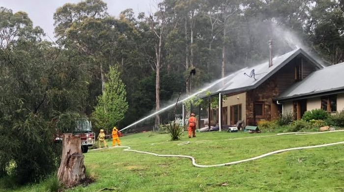 Firefighters spray a hose at the roof of a house. Trees in background.
