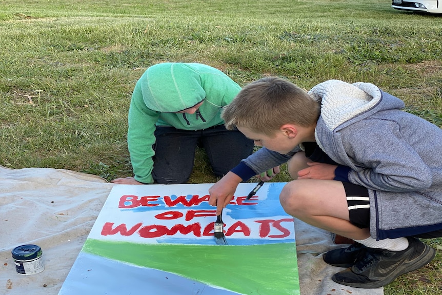 Two boys crouch down over their half-painted wombat road sign and continue painting.