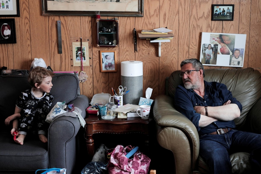 An young boy and an older man are pictured sitting on two different sofas, talking to each other. On the wall framed pictures