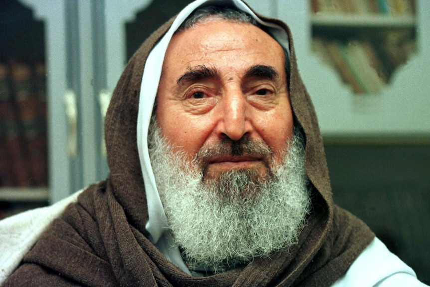 An old man with white beard sits and looks into camera, wearing brown and white head covering
