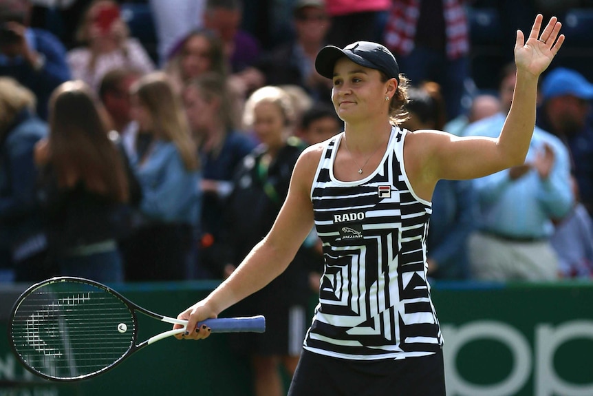 Ash Barty smiling and holding her tennis racquet in her right hand and waving with her left.